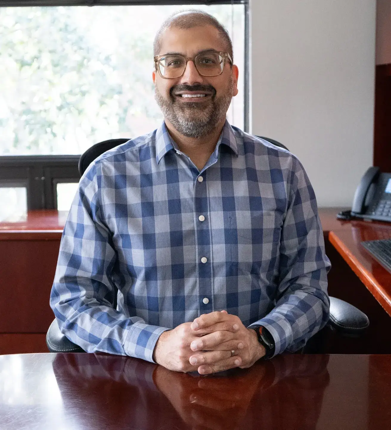 Man in glasses and plaid shirt sitting at desk, smiling.